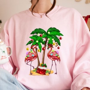 May include: Pink sweatshirt featuring a festive design with two flamingoes adorned with Christmas lights and ornaments, standing under palm trees decorated with Christmas lights and ornaments. A red gift bag is at the base of the trees. The sweatshirt is a solid pink color.