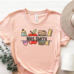 May include: A peach-colored t-shirt with a graphic design of a red apple, a pencil, a coffee cup, and a ruler. The text "MRS. SMITH" is printed on the shirt.