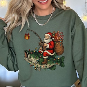 May include: An olive green sweatshirt features a whimsical Christmas design. Santa Claus, dressed in his traditional red suit, is depicted fishing while riding a bass decorated with Christmas lights. A sack of candy canes and a wrapped gift complete the festive scene.