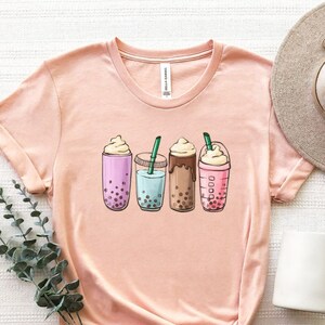 May include: A pink t-shirt with a graphic of four different bubble tea drinks. The drinks are all in clear cups with colorful lids and straws. The graphic is in a cartoon style.