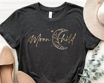 May include: Dark gray t-shirt with the words "Moon Child" in gold script, accompanied by a crescent moon design. The shirt is styled with tan loafers and a wide-brimmed hat, with sprigs of greenery.