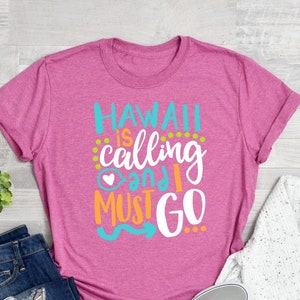 Może przedstawiać: Różowa koszulka z napisem "HAWAII is calling and I MUST GO" w białym, niebieskim i pomarańczowym kolorze. Koszulka leży na drewnianej powierzchni z dżinsami i białym swetrem.