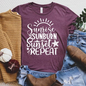 Könnte beinhalten: Ein maroonfarbenes T-Shirt mit weißem Text, der "Sunrise Sunburn Sunset Repeat" lautet, mit einem Seestern-Motiv. Das Shirt liegt auf einer weißen Oberfläche mit einem braunen Pullover, einer blauen Jeans und braunen Stiefeln.
