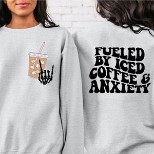 Könnte beinhalten: Grauer Pullover mit einem Grafikdruck eines Glases Eiskaffees und einer Skeletthand auf der Vorderseite. Die Rückseite des Pullovers trägt den Text "Fueled by Iced Coffee & Anxiety" in Schwarz.