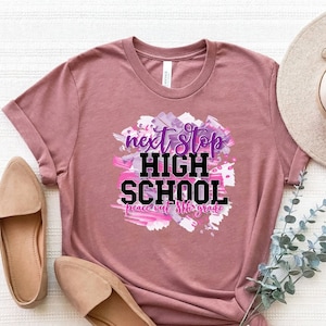 May include: A mauve t-shirt with a graphic design that says "next stop high school peace out 8th grade" in black and purple text on a pink and purple watercolour background.