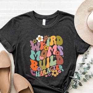 May include: A dark grey t-shirt with a colourful retro-style graphic that says "Weird Moms Build Character". The graphic features flowers and a heart.