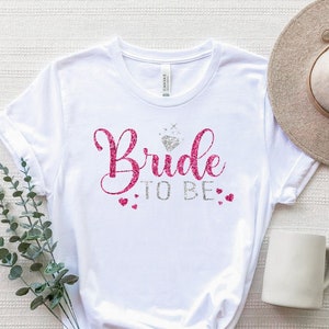 May include: White t-shirt with pink glitter text that says "Bride to Be" with a silver glitter diamond and small pink hearts.