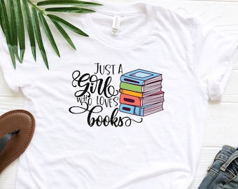 Può includere: Maglietta bianca con il testo "Just a girl who loves books" in corsivo nero. Accanto al testo è illustrata una pila di libri colorati.