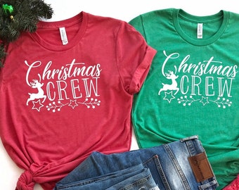 May include: Two Christmas themed t-shirts with the text "Christmas Crew" and a reindeer graphic. The shirts are red and green and are laid out on a white background with a pair of blue jeans and a red beanie.