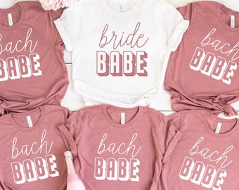 Może przedstawiać: Sześć różowych koszulek z białym nadrukiem. Koszulki mają napis "bach babe" i "bride babe".