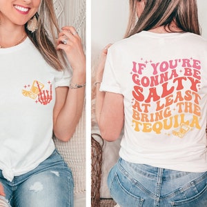 May include: White t-shirt with a front pocket design featuring a skeleton hand, a cocktail, and lemon slices in orange and yellow. The back of the shirt displays the text "IF YOU'RE GONNA BE SALTY AT LEAST BRING THE TEQUILA" in a retro font.