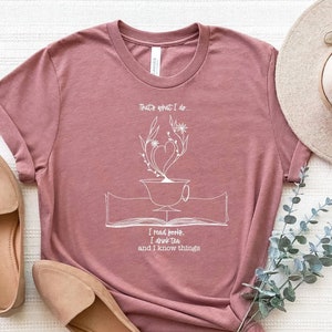 May include: A mauve t-shirt with a white graphic design of a teacup with a heart-shaped steam rising from it, surrounded by leaves and flowers. The text "That's what I do. I read books, I drink tea and I know things" is printed below the graphic.