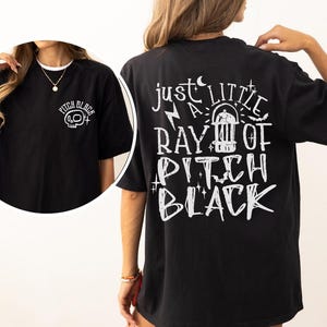 May include: Black t-shirt with white graphic text on the back that reads "just a little ray of pitch black." The front of the shirt has a small graphic of a skull and the words "pitch black."