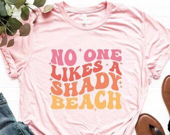 Puede incluir: Camiseta rosa con el texto "NO ONE LIKES A SHADY BEACH" en una fuente retro de colores arcoíris.