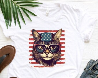May include: White t-shirt featuring a cat wearing sunglasses and a hat with stars, set against a backdrop of red and white stripes. The cat graphic has a vintage, patriotic design. The shirt is paired with blue jeans and black sandals.