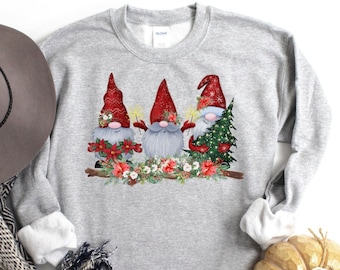 May include: A gray sweatshirt with a Christmas gnome design. Three gnomes are holding sparklers and a Christmas tree. The gnomes are wearing red hats and have white beards. The design is surrounded by red poinsettias and green foliage.