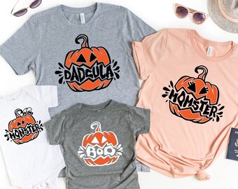 Puede incluir: Un conjunto de cuatro camisetas con temática de Halloween con un diseño de calabaza. Las camisetas son de color gris, melocotón y blanco. El texto en las camisetas dice "Dadcula", "Momster", "Mini Monster" y "Boo".