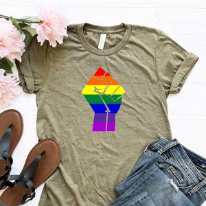 May include: A green t-shirt with a rainbow fist graphic. The fist is raised in the air and the colors of the rainbow are in the order of the pride flag. The shirt is laying on a white surface with a pair of black sandals and blue jeans.
