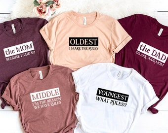 Puede incluir: Cinco camisetas de manga corta en varios colores: burdeos, melocotón, malva y blanco. Cada camiseta tiene un diseño de texto diferente en un rectángulo negro: "the MOM BECAUSE I SAID SO", "OLDEST I MAKE THE RULES", "the DAD GO ASK YOUR MOM", "MIDDLE I'M THE REASON WE HAVE RULES" y "YOUNGEST WHAT RULES?"