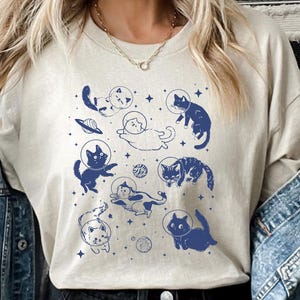 May include: A beige t-shirt with a graphic print of cats in space. The cats are wearing astronaut helmets and floating around planets. The graphic is in shades of blue.