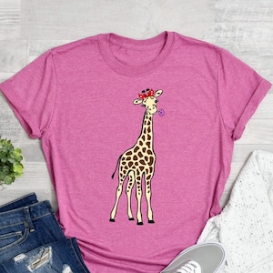 May include: A pink t-shirt featuring a cartoon giraffe wearing a red bandana with white polka dots and a purple flower. The giraffe is blowing a purple flower.