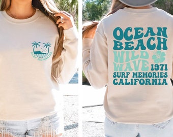 May include: A light beige sweatshirt with a teal blue graphic of palm trees and waves on the front and the text "Ocean Beach Wild Wave 1971 Surf Memories California" on the back.