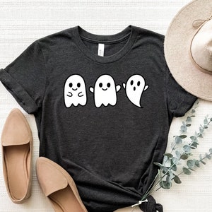 May include: A dark gray t-shirt with a graphic of four white ghosts. The ghosts have different expressions, including a smiling ghost, a ghost with its arms crossed, and a ghost with its mouth open.