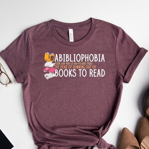 May include: A maroon t-shirt with a graphic design that says "ABIBLIOPHOBIA" and "THE FEAR OF RUNNING OUT OF BOOKS TO READ".