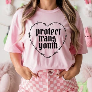 以下が含まれることがあります： 「protect trans youth」の文字が、ハート型の有刺鉄線デザインの中に描かれた、薄いピンク色のTシャツ。ピンクと白のチェック柄のパンツを着用。背景にはイースターの飾りがあります。