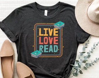 May include: A dark gray t-shirt with the text "LIVE LOVE READ" in a colorful retro-style font. The text is surrounded by a colorful frame and stars. There are stacks of books on either side of the text.