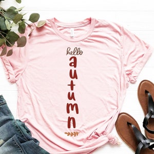 Peut inclure: Un t-shirt rose avec le texte "hello autumn" imprimé verticalement en lettres brunes. Le t-shirt est associé à un jean bleu et des sandales brunes.