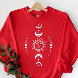 Puede incluir: Sudadera roja con un dise&ntilde;o blanco de sol y luna. El sol tiene una cara y est&aacute; rodeado por un c&iacute;rculo de lunas.