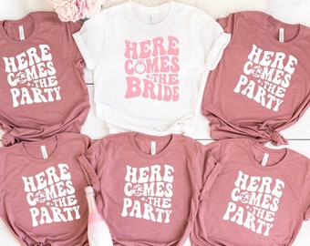Puede incluir: Una colección de camisetas de manga corta en color malva y blanco, con las frases "Here Comes the Party" y "Here Comes the Bride" en una fuente retro. También se ve una botella de champán.