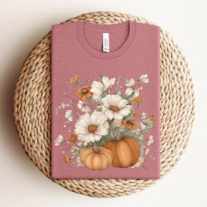 May include: A dusty rose t-shirt featuring a floral design with white and orange flowers, and two pumpkins. The shirt is folded on a woven, circular placemat. The tag on the shirt is white.