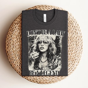 Op de afbeelding: Donkergrijs t-shirt met een zwart-wit afbeelding van een vrouw met een zonnebril. De tekst "DON'T BE A LADY BE A LEGEND" staat boven en onder de afbeelding. Het shirt wordt getoond op een geweven, cirkelvormig oppervlak.