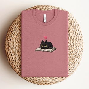 以下が含まれることがあります： 黒猫が本の上に寝そべり、ピンクのハートが描かれた刺繍デザインが特徴のダスティローズTシャツ。Tシャツはクルーネックで、背面上部にタグが付いています。