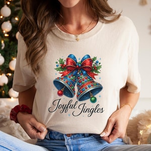 Può includere: T-shirt color crema con un design festivo di due campane natalizie blu adornate con fiocchi rossi, agrifoglio e ornamenti colorati. Le parole "Joyful Jingles" sono scritte in un carattere corsivo sotto le campane.