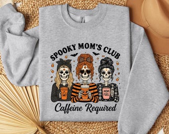 May include: A light gray sweatshirt with the text "Spooky Mom's Club" and the phrase "Caffeine Required." The graphic features three skeleton women holding coffee cups. The women have different hairstyles and wear bows and patterned tops.