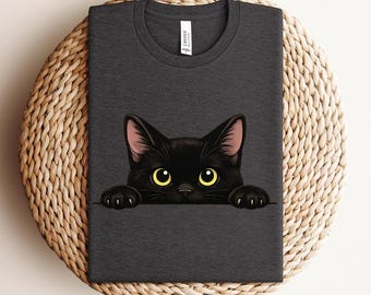 Puede incluir: Camiseta gris oscuro con un gráfico de gato negro con ojos amarillos y orejas rosas. La cabeza y las patas del gato asoman por encima de una línea gris oscuro. La camiseta tiene cuello redondo y una pequeña etiqueta blanca.