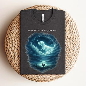 Puede incluir: Camiseta gris oscuro con cuello redondo. Presenta una ilustración surrealista de un dinosaurio brillante dentro de un óvalo lleno de nubes, con una luna creciente y una silueta de pollo. El texto "remember who you are." está encima de la ilustración.