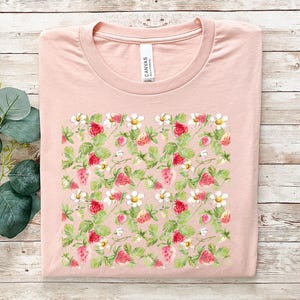 Könnte beinhalten: Ein pinkfarbenes T-Shirt mit einem weißen und grünen Erdbeer-Druck. Die Erdbeeren sind rot und haben grüne Blätter. Das Shirt hat einen Rundhalsausschnitt und kurze Ärmel.