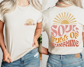 May include: Cream-colored t-shirt with the word "SUNSHINE" and a sun graphic on the front. The back of the shirt features the text "SOUL FULLS OF SUNSHINE" with a sunburst design. The shirt is paired with blue denim jeans.