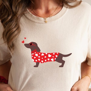 Puede incluir: Una camiseta blanca con un dachshund marr&oacute;n que lleva gafas de sol en forma de coraz&oacute;n rojo y un su&eacute;ter rojo con corazones blancos. El dachshund mira hacia la izquierda y lanza un beso.