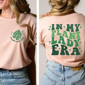 May include: A pink t-shirt with a green leaf graphic and the text "Plant Lady Era" on the front. The back of the shirt has the text "In My Plant Lady Era" in green letters with stars.