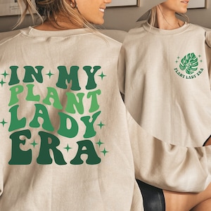 May include: A beige sweatshirt with the text "In My Plant Lady Era" in green and white on the back. The front of the sweatshirt has a green leaf with the text "Plant Lady Era" in green and white.