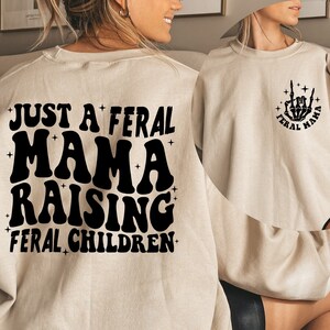 May include: Two beige sweatshirts with black text and graphics. One sweatshirt says "Just a Feral Mama Raising Feral Children". The other sweatshirt has a skeleton hand making a rock and roll sign with the words "Feral Mama" around it.