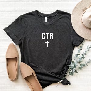 Op de afbeelding: Donkergrijs t-shirt met de witte tekst "CTR" en een kruissymbool. Het shirt wordt gecombineerd met beige instappers en een breedgerande hoed. Een takje groen voegt een decoratief tintje toe.