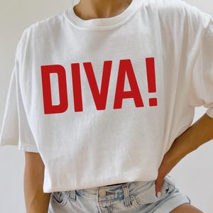 May include: A white short-sleeved t-shirt featuring the word DIVA! printed in bold red capital letters across the chest. The garment has a crew neckline and a relaxed fit, paired with light blue denim shorts with a visible metal button and rivets.