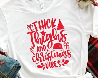 Puede incluir: Camiseta blanca con texto rojo que dice "Thick Thighs and Christmas Vibes" con gráficos de árbol de Navidad, bastón de caramelo, gorro de Papá Noel y caja de regalo.