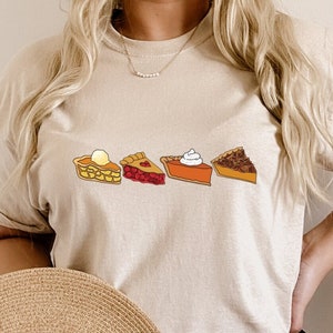 Puede incluir: Una camiseta beige con un gráfico de cinco rebanadas de pastel diferentes. Las rebanadas son un pastel de nuez pecana, un pastel de calabaza, un pastel de cereza, un pastel de manzana y un pastel de melocotón. La camiseta la lleva puesta una persona con el pelo largo y rubio.